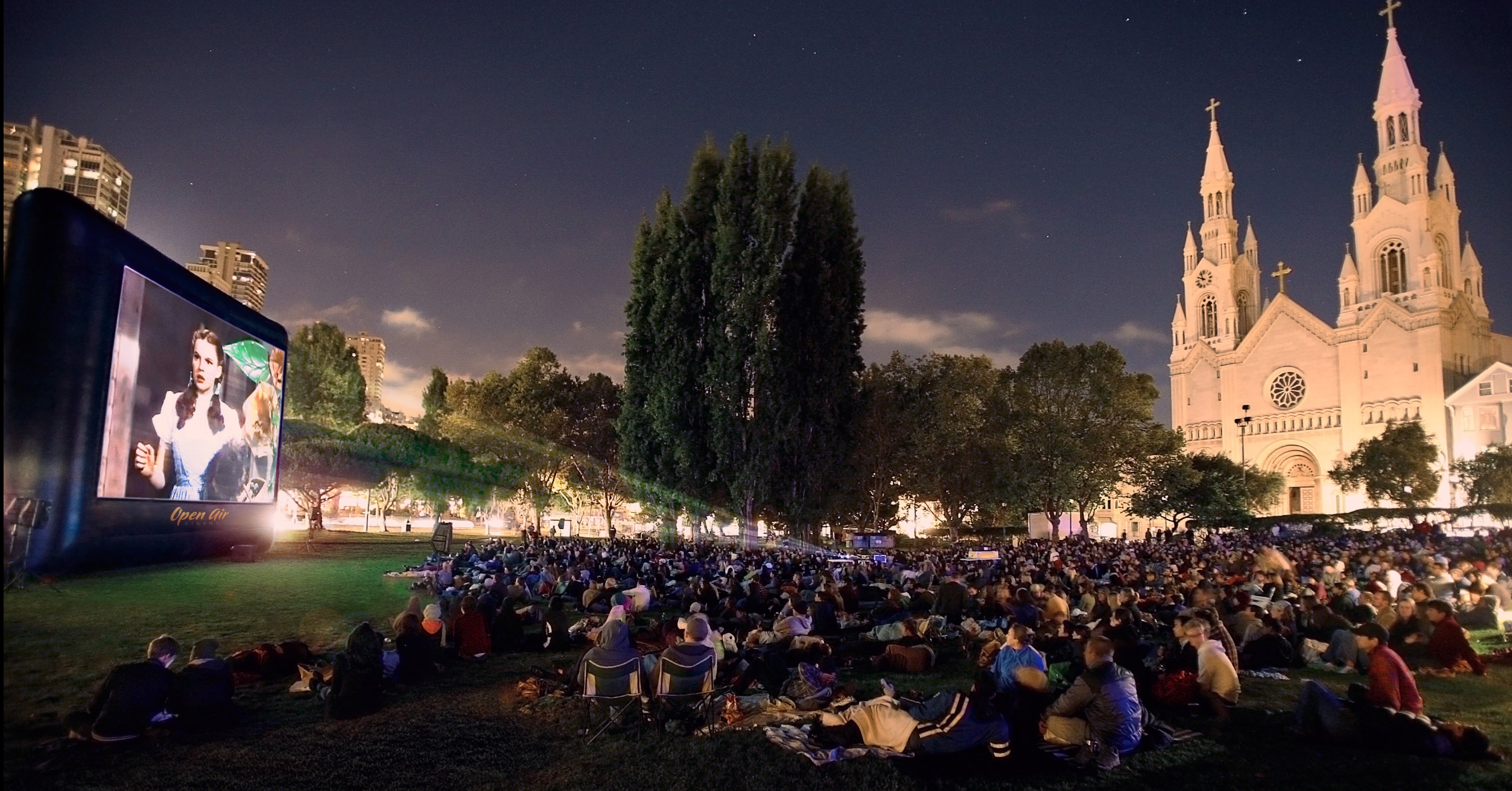 A large night-time audience seated on the grass in front of a lit Open Air Cinema screen
